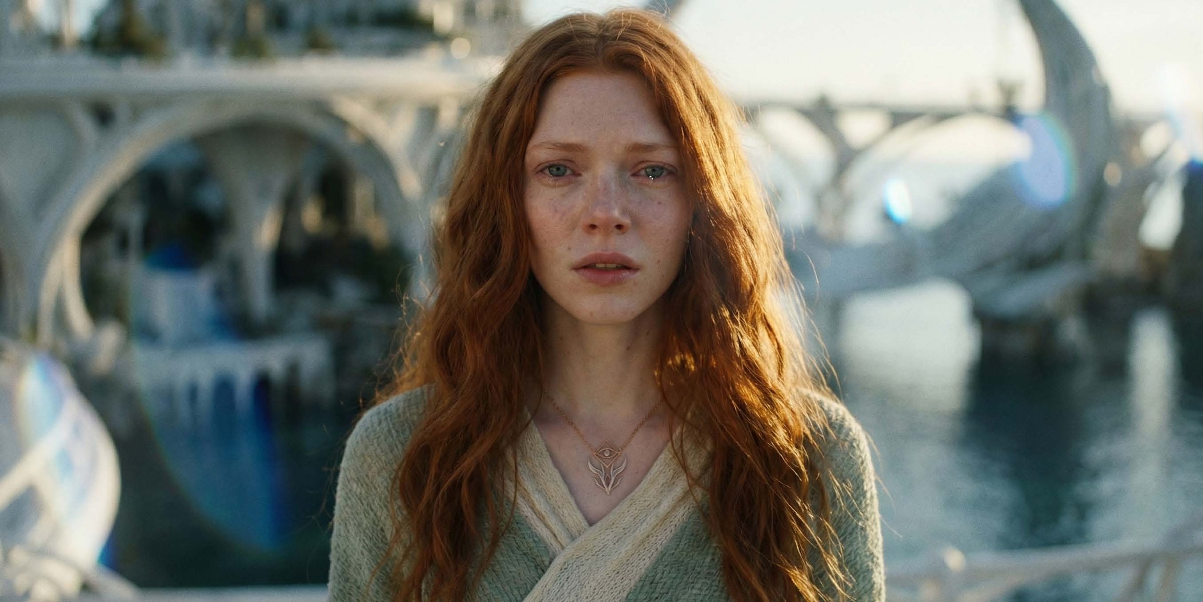 A young woman with long, wavy copper-red hair and freckles looks directly at camera, lips slightly parted and her eyes wet with tears. She wears a pale sage and cream knitted wrap sweater and a fine gold chain necklace w
