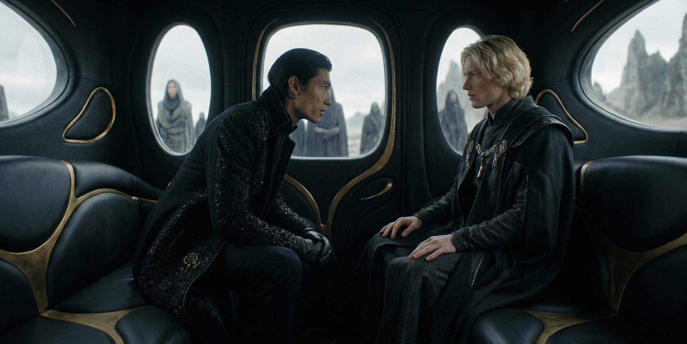 Interior of a sleek dark vehicle or shuttle with smooth padded black leather seats divided by sculpted brass-gold inlay, and oval windows rimmed in gold. On the left a dark-haired man in an ornate black brocade coat lean