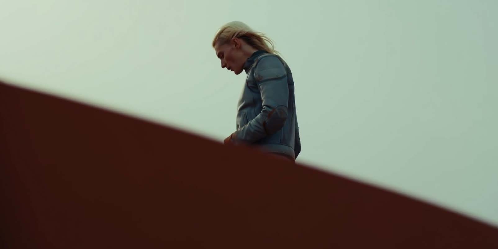 Low-angle cinematic still: a blonde woman in a pale blue-grey utility jacket stands at the top of a steep dark red-brown wall or embankment, head bowed and hands together in front of her, lit against a flat pale mint-gre