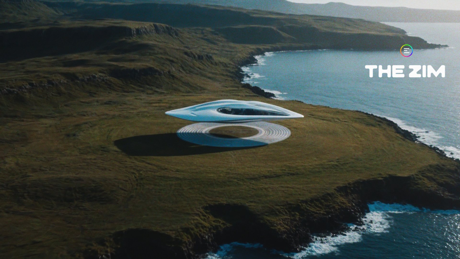 Aerial view of a low, white ring-shaped structure set on a green coastal headland above the sea.