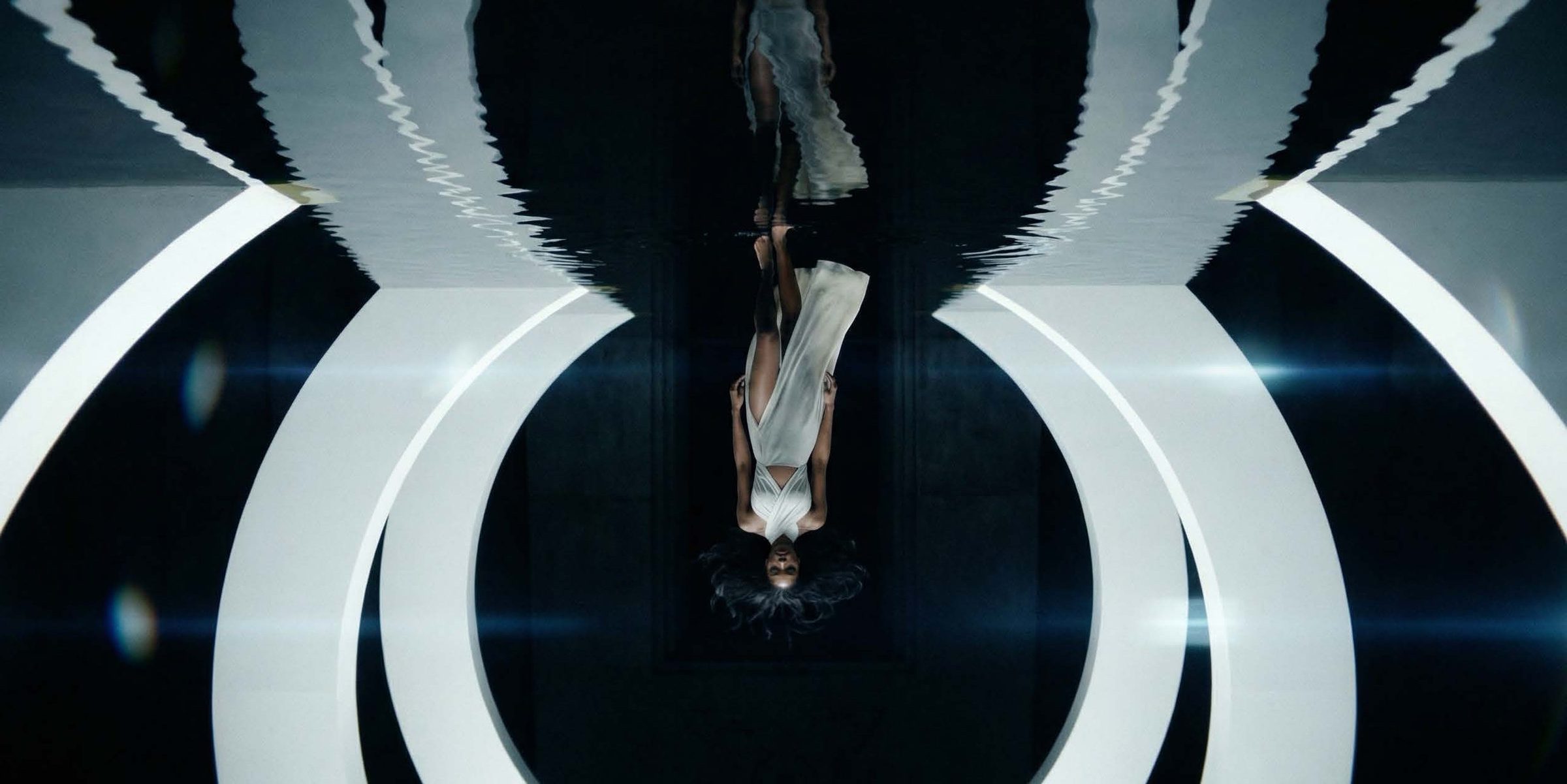 A woman in a pale draped gown with a high thigh slit walks barefoot toward camera down a dark corridor, her long hair floating around her head, flanked by tall curved white light arcs and mirrored in the sheet of black w